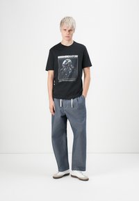 Young person with short blond hair wearing black graphic T-shirt reading "Synthetic Nature," loose blue pants with white drawstrings, and white sneakers.