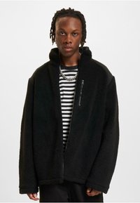 Schwarze Fleecejacke mit hohem Kragen, Reißverschlusstasche auf der Brust und weiten Ärmeln, kombiniert mit einem schwarz-weiß gestreiften Hemd darunter.