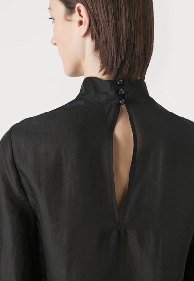 Blusa nera con colletto alto, chiusura con bottoni sul retro e fessura sotto il colletto. Il tessuto ha una texture liscia e leggermente lucida.