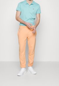 Hellblaues Polohemd, orangefarbene Sweatpants und weiße Turnschuhe. Das Polohemd hat ein kleines Logo; die Hose hat einen elastischen Bund und umschlossene Knöchel.