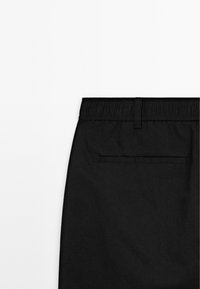 Pantaloni neri con una cintura elastica, dotati di una tasca sul retro. Il tessuto appare liscio su uno sfondo bianco semplice.