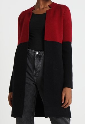 Femme portant un long cardigan ouvert devant, avec une moitié supérieure rouge et une moitié inférieure noire, assortie à un haut noir et un jean foncé.