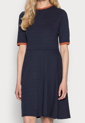 Frau trägt ein marineblaues, geripptes, knielanges Kleid mit kurzen Ärmeln und orangefarbenen Akzenten am Kragen und an den Ärmelsäumen.