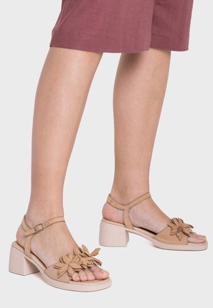 Kobieta nosząca beżowe sandały na klocku z ozdobami kwiatowymi i paskami wokół kostki, połączone z mauve spódnicą do kolan.