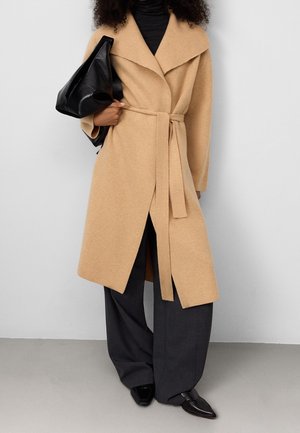 Personne portant un manteau en laine beige ceinturé sur un pantalon noir et un haut noir, tenant un grand sac à main en cuir noir sur une épaule.
