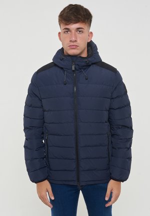 Giacca puffer blu navy con design trapuntato, cerniere laterali e cappuccio. Caratterizzata da dettagli neri sulle spalle e sui polsini. Materiale leggero e isolato.
