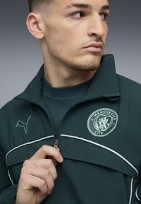Grüne Jacke mit weißen Akzenten, ausgestattet mit einem Manchester City-Logo und einem Puma-Wappen. Mit Reißverschluss vorne und hohem Kragen.
