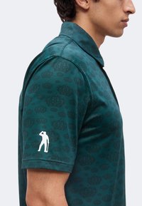 Homme portant un polo bleu canard avec un motif de couronne subtil et un petit logo blanc en silhouette d'une personne tenant un club de golf sur la manche.