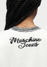 Άτομο με μαύρα στριφτά μαλλιά, φωτογραφημένο από πίσω, φοράει λευκό πουλόβερ με την επιγραφή "Moschino Jeans" κεντημένη με μαύρα γράμματα στην πλάτη.