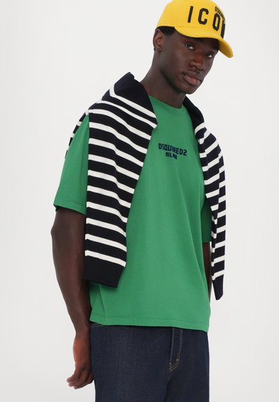 Grønn bomull t-skjorte med svart brodert logo, kombinert med enstripete marineblå og hvit genser lagt over skuldrene og mørke jeans.