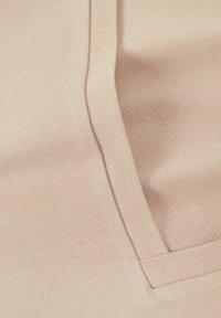 Beige tyg med en slät textur, med V-halsringning och sydda detaljer. Synliga sömmar ger struktur åt den övergripande designen.
