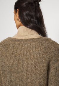 Beige trui met hoge hals en een geribbelde textuur, met een losse pasvorm en een zachte, pluizige afwerking. Subtiele glans en lichtbruine accenten.