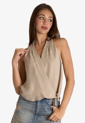 Giovane donna con lunghi capelli castani che indossa una canottiera beige senza maniche a fascia incrociata e jeans blu usurati, guarda di lato con la mano vicino al colletto.