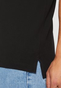 Svart texturerad t-shirt med insida split detalj, som visar ljusblå denimshorts längst ner. Närbild av tyg och sömnad.