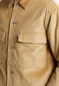 Beige cordjacka i manchester med en texturerad, ribbad yta, två bröstfickor och silverfärgade tryckknappar. Har en mjuk, kragliknande halsringning.