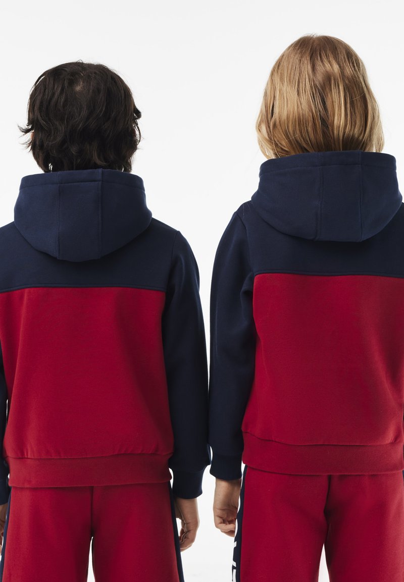 Zwei Kinder stehen Rücken an Rücken und wenden sich dem Betrachter zu. Sie tragen passende rote und marineblaue Kapuzenpullover sowie rote Hosen.