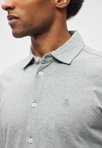 Polo cinzento com textura suave, gola curta, vista com três botões e logo bordado subtil no peito.