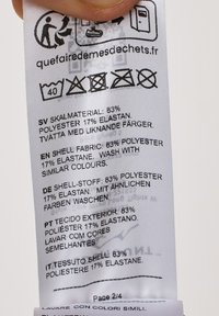 Vaskeanvisning på tekstilmærke med flere sprog, der viser tekstilmaterialets sammensætning: 83% polyester, 17% elastan; vaskeanvisninger angivet med symboler.