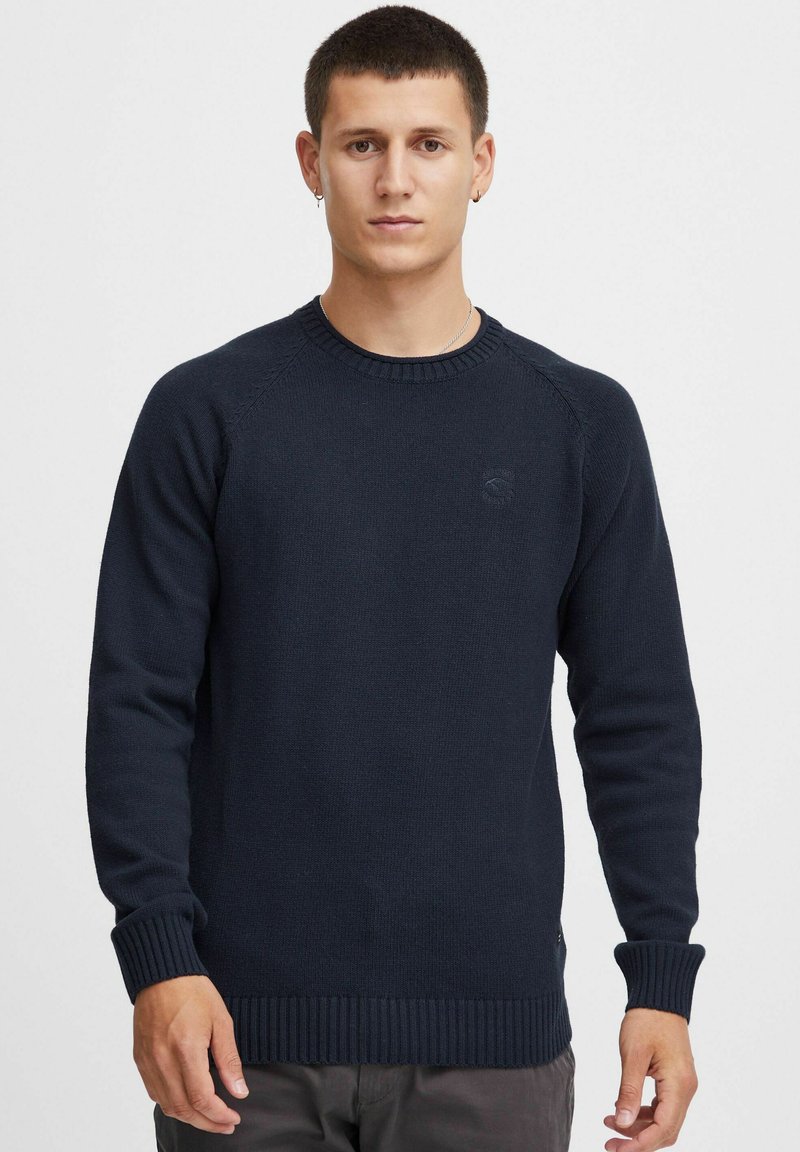 Navy blue knitted sweater with ribbed cuffs and hem, featuring a round neckline and subtle logo near the chest. Standard fit and texture.