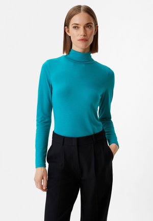Türkiser Rollkragenpullover mit langen Ärmeln aus glattem Stoff, enganliegendes Design, kombiniert mit schwarzen Hosen mit maßgeschneidertem Schnitt und Taschen.