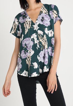 Femme portant un chemisier vert à manches courtes avec un motif floral violet et blanc, associé à un pantalon noir, debout devant un fond uni.