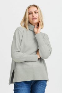 Un pull à col roulé gris avec une texture côtelée et des fentes latérales, fabriqué à partir d'un tissu doux. Manches longues et coupe décontractée, associé à un jean bleu.