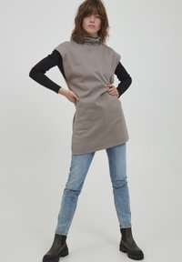 Prevelik bež brezrokavnik s turtleneck iz mehke tkanine z rebrasto teksturo, v kombinaciji z oprijetimi svetlo modrimi kavbojkami in črnimi debelimi gležnjarji.