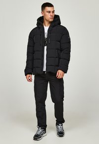 Schwarze Pufferjacke mit Kapuze, ausgestattet mit einem gesteppten Design, Reißverschluss vorne und verstellbaren Zügen. Kombination mit passenden schwarzen Cargo-Hosen und Sneakers.