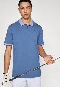 Μπλε πόλο μπλούζα με κολάρο με σχέδιο, κοντά μανίκια και μικρό λογότυπο στο στήθος. Φορεμένη από άτομο που κρατά ένα γκολφ κλαμπ.