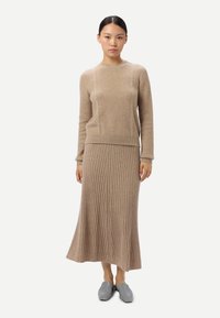 Femme portant un pull en tricot beige et une jupe midi côtelée assortie, associée à des chaussures grises faciles à enfiler, debout devant un fond blanc uni.