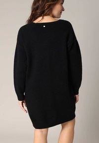 Robe tricotée noire, oversized, avec un ourlet et des poignets côtelés. Présente un décolleté dos bas et un petit détail doré au dos.