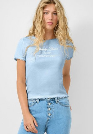 Junge Frau mit langen, lockigen blonden Haaren, die ein hellblaues, tailliertes T-Shirt und eine hoch taillierte blaue Jeans mit sichtbaren Knöpfen trägt.