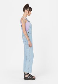 Salopette in denim azzurro chiaro con spalline regolabili, tasche laterali e gamba dritta, abbinata a una canotta lavanda e sandali neri.