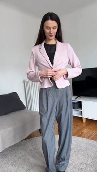 Žena zakopčava svijetloružičasti sako preko crnog topa, nosi lepršave sive hlače i stoji u modernom dnevnom boravku.