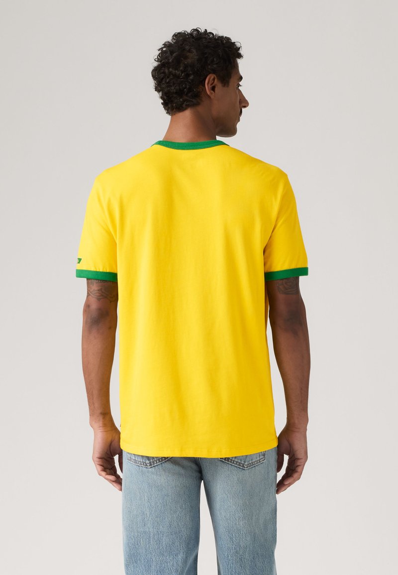 Homme aux cheveux bouclés portant une chemise jaune à manches courtes avec un bord vert, debout de dos, portant un jean bleu clair sur un fond uni.