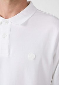 Witte polo shirt gemaakt van textuurstof, voorzien van een klassieke kraag en drie knopen aan de voorkant. Opvallende witte logo patch op de linkerkant van de borst.
