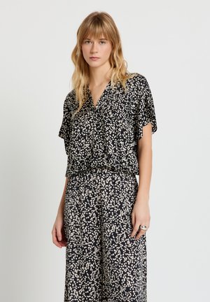 Giovane donna con capelli biondi che indossa una camicetta a maniche corte e pantaloni a gamba larga coordinati con motivo bianco e nero, in piedi davanti a uno sfondo semplice.