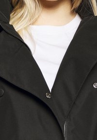 Primo piano di una persona che indossa una camicia bianca e un giubbotto nero con una cerniera parzialmente aperta e bottoni visibili.