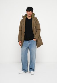 Olivengrøn polstret jakke med lynlås, snørehætte og trykknapper. Båret over en sort sweater og lyseblå bredbenede jeans med hvide sko.