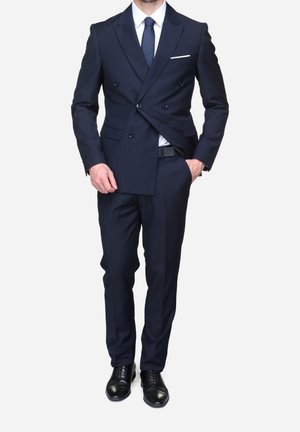 Homme portant un costume bleu marine croisé sur mesure, chemise blanche, cravate bleu marine, chaussures habillées noires, une main dans la poche et l'autre ajustant la veste.