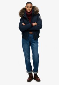 Marineblå pufferjakke med pelsforet hætte, bordeaux turtleneck og blå denimjeans. Brune snørebodstøvler med opfoldede støvler.