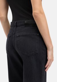 Zwarte spijkerbroek met een hoge taille, voorzien van achterzakken en een leren label. De stof heeft een gladde textuur en een smalle pasvorm.