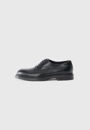 Chaussure en cuir noir avec une finition texturée, bout rond, cinq œillets et une semelle basse. Présente un logo discret au niveau du talon.
