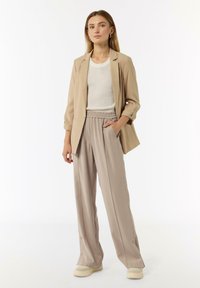 Beige blazer over a white knit top, paired with light beige pleated trousers. Soft fabric, loose fit, subtle sheen, slip-on shoes.