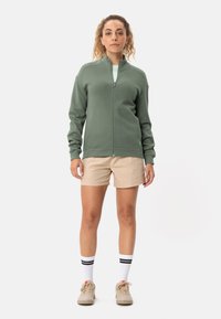 Giacca in pile verde con zip e un motivo testurizzato, indossata sopra una camicia chiara, abbinata a pantaloni corti beige e calze sportive bianche con strisce nere.