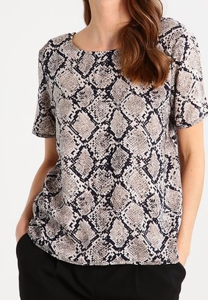 Kobieta w bluzce z krótkim rękawem o beżowo-czarnym wzorze w skórę węża, w czarnych spodniach, z rękami w kieszeniach.