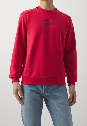 Uomo con indosso una felpa rossa semplice con un piccolo logo nero EA7 sul petto e jeans azzurri, in piedi contro uno sfondo bianco.