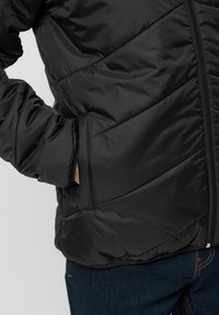 Veste matelassée noire avec un design en losanges, fermeture éclair et poche latérale. Fabriquée en matériau lisse et léger, montrée de profil.