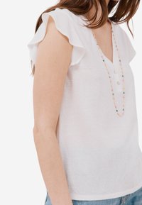 Top bianco in lino con maniche corte arricciate, scollatura a V, chiusura frontale con tre bottoni e una collana colorata di perline che esalta il design.