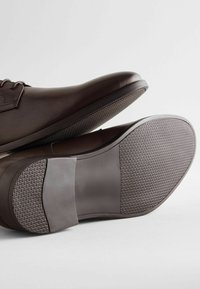Zapatos de cuero marrón con punta redondeada, diseño con cordones y suela de goma texturada. Presentan una parte superior lisa y un patrón de tracción contrastante.